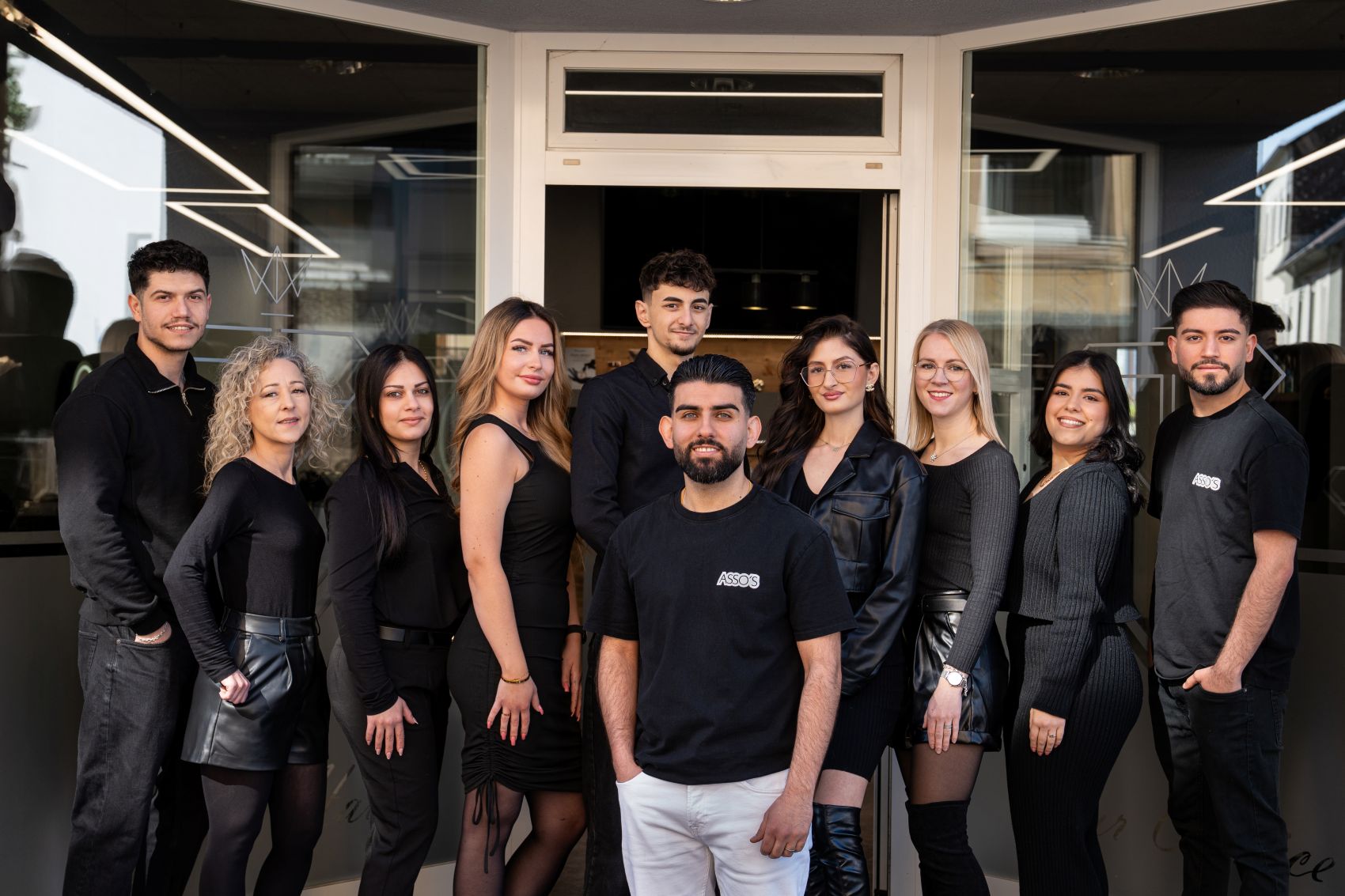Teambild von Assos Hairdesign in Bitburg vor dem Friseursalon-Eingang. Es sind insgesamt 4 Friseure und 6 Friseurinnen auf dem Bild zu sehen.